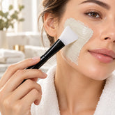 A woman applies a light green face mask to her cheek with a black and white silicone brush. Her hair is tied back, and she