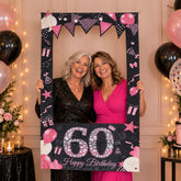 Two smiling women holding up a black and pink 60th birthday photo booth frame with balloons and gifts design.