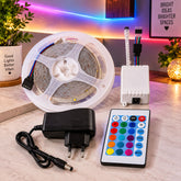 A coiled RGB LED strip light, white controller, power adapter, and remote control on a wooden surface.