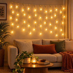 Warm white bulb string lights hang on a wall above a sofa with pillows, a plant, and a coffee table with a candle.
