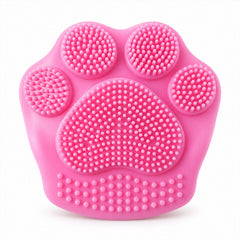 A bright pink paw-shaped facial scrubber with numerous silicone bristles is centered on a white background.