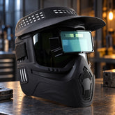 A black welding helmet with a visor and green-tinted auto-darkening lens sits on a metal workbench in a workshop.