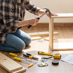A person in a plaid shirt and jeans kneels on a wooden floor, using a hammer to assemble a light wood frame.