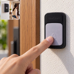 A person's finger presses the light grey button of a black wireless doorbell transmitter on a white wall next to a wooden