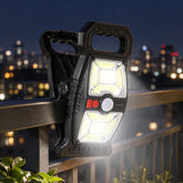 A black solar motion sensor clip light illuminates a dark city balcony railing at night, with distant buildings blurred.