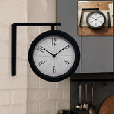 A black double-sided wall clock with a white face and black numbers is mounted on a white brick wall in a kitchen setting. A