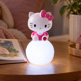 A Hello Kitty LED night light illuminates a wooden table next to a children's book and a potted plant.