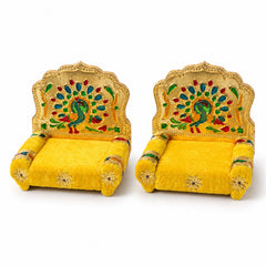 A pair of golden singhasans with yellow velvet upholstery and peacock-decorated backrests, set against a plain white