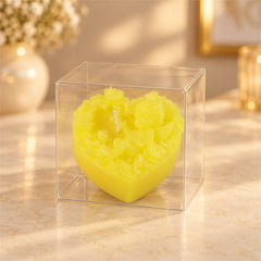 A vibrant yellow heart-shaped candle with intricate floral and cherub details, encased in a clear plastic box.