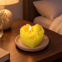 A lit yellow heart-shaped candle with a cherub and floral motif, sitting on a small plate on a bedside table.