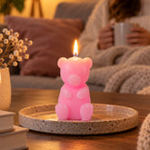 A lit pink teddy bear candle sits on a speckled ceramic plate, with a person holding a mug in the background.