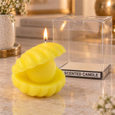 A lit yellow shell-shaped candle with a pearl center sits beside its clear plastic packaging on a marble surface.