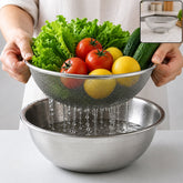 Hands hold a stainless steel mesh strainer full of fresh vegetables and lemons over a steel bowl of water