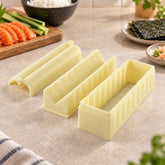 A three-piece light yellow plastic sushi maker set on a light countertop, with ingredients in the background.