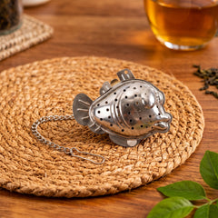 A metallic fish-shaped tea infuser with a chain and hook rests on a round woven placemat on a wooden surface.