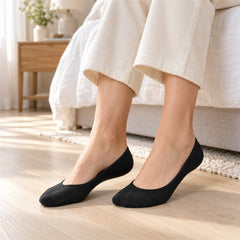 A person's feet wearing black low-cut socks, sitting on a light wooden floor beside a bed in a bright room.
