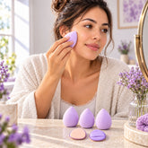 A woman applies makeup with a lavender blending sponge, with three sponges and two puffs on a table.