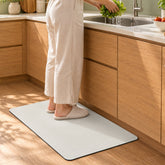 A person in slippers stands on a cream rectangular floor mat in a modern kitchen with wooden cabinets. They are washing