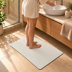 A person in a robe stands barefoot on a cream mat in a bathroom, washing hands at a white basin.