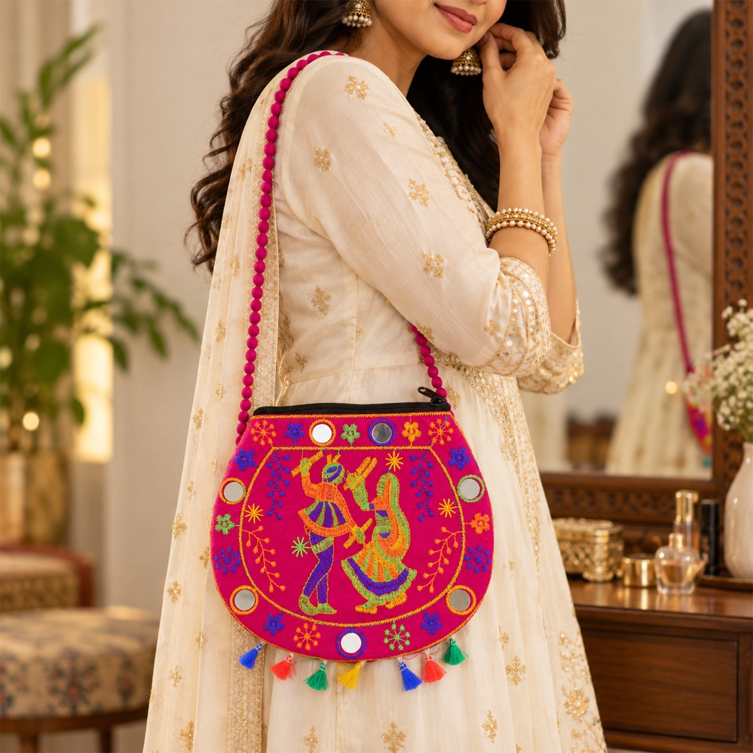 A woman in a cream dress wears a vibrant pink sling bag with embroidered dancers and small mirrors.