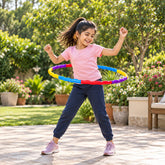 A joyful girl in a pink shirt and blue pants spins a red, blue, yellow, and purple hula hoop outdoors.