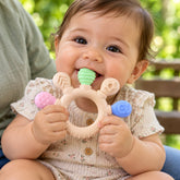 A happy baby in a floral onesie chews on a beige teether with green, pink, and blue textured ends.