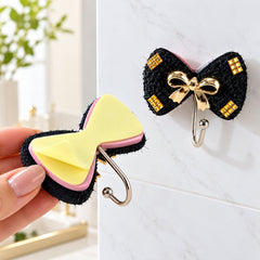 A hand holding one black bow-shaped hook with a yellow adhesive backing, next to another hook mounted on a white wall.