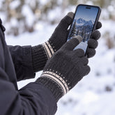 Hands wearing dark gray knitted gloves with white patterned cuffs using a smartphone in a snowy outdoor setting.