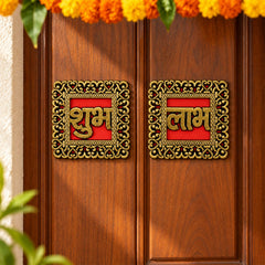 Two square Shubh Labh decals with intricate gold borders on a rich brown wooden door, sunlight casting a shadow.