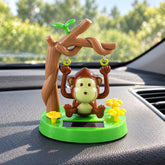 A brown monkey figure swings from a tree branch on a green base, placed on a car dashboard with a blurred outdoor background.
