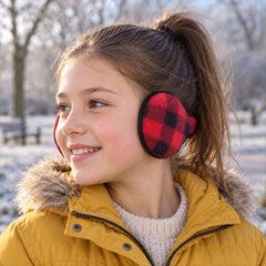 A young girl with a ponytail wears red and black plaid ear warmers in a snowy park setting.