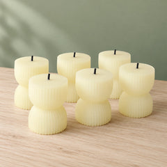 Six unlit cream-colored ribbed LED candles with black wicks arranged on a light wooden surface against a green wall.