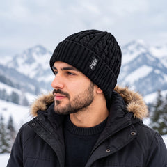 A man wearing a black cable knit beanie with a ribbed cuff and a small brand label, against a snowy mountain backdrop.