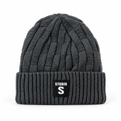 A dark grey cable knit beanie with a ribbed cuff and a black label with "STUDIO S" in white, against a plain white
