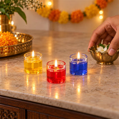 Three lit gel candles in yellow, red, and blue sit on a counter with a hand placing a white flower in a brass bowl.