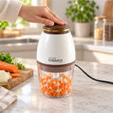 A hand presses the brown top of a white and clear electric chopper filled with diced carrots and onions on a kitchen counter.