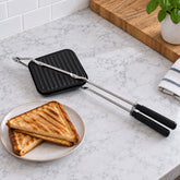 Black stovetop sandwich maker with long metal handles and two golden brown grilled sandwiches on a white plate.