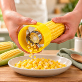 Hands using a yellow corn stripper, removing kernels from a cob into a white bowl on a wooden surface.