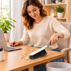 A person uses the white and gray brush to sweep crumbs from a wooden desk into a waste bin.