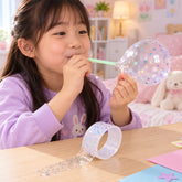 A young girl blows into a light green straw, inflating a clear glitter-flecked bubble from craft tape.