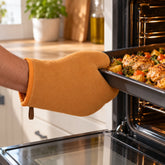 A hand wearing an orange oven mitt pulls a baking tray with roasted chicken and vegetables from a hot oven.