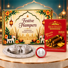 A festive gift hamper on a light brown pedestal, featuring a colorful box, red wafer rolls, a silver puja thali, and a small