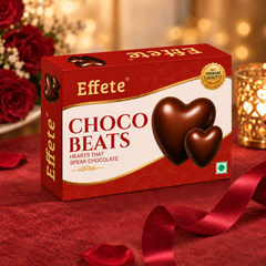 A box of Effete Choco Beats chocolates is surrounded by red roses, petals, a ribbon, and a glowing candle.