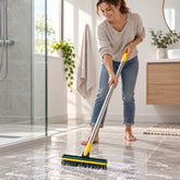 A woman in jeans and a sweater scrubs a tiled bathroom floor with a green and yellow brush, creating suds.