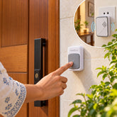 A person's hand presses a gray and white wireless doorbell button mounted on a light-colored wall next to a wooden door.