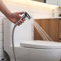 A hand holds a chrome bidet sprayer, spraying water towards a white toilet in a tiled bathroom with warm lighting.