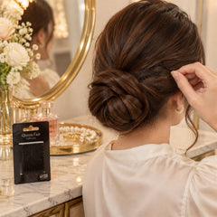 A woman uses a black bobby pin to style her hair bun, with a mirror and perfume bottle in the background.