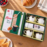 Open gift box revealing a white bottle and four white ceramic mugs with black speckled rims on a wooden table.