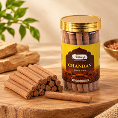 A clear jar of brown dhoop sticks stands on a wooden board, with a pile of sticks in front.