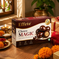 A box of Effete Choco Magic chocolates on a light wooden table with marigold flowers, a lit candle, and a golden gift.